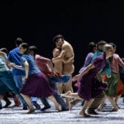 A group of dancers in colorful clothing move energetically on stage while two dancers, one in nude attire, embrace at the center.