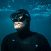 A person wearing a black wetsuit, swim cap, and goggles is underwater, looking upward with light filtering through the water above—capturing the adventurous spirit of the Ocean Film Festival.