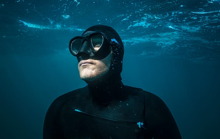 A person wearing a black wetsuit, swim cap, and goggles is underwater, looking upward with light filtering through the water above—capturing the adventurous spirit of the Ocean Film Festival.