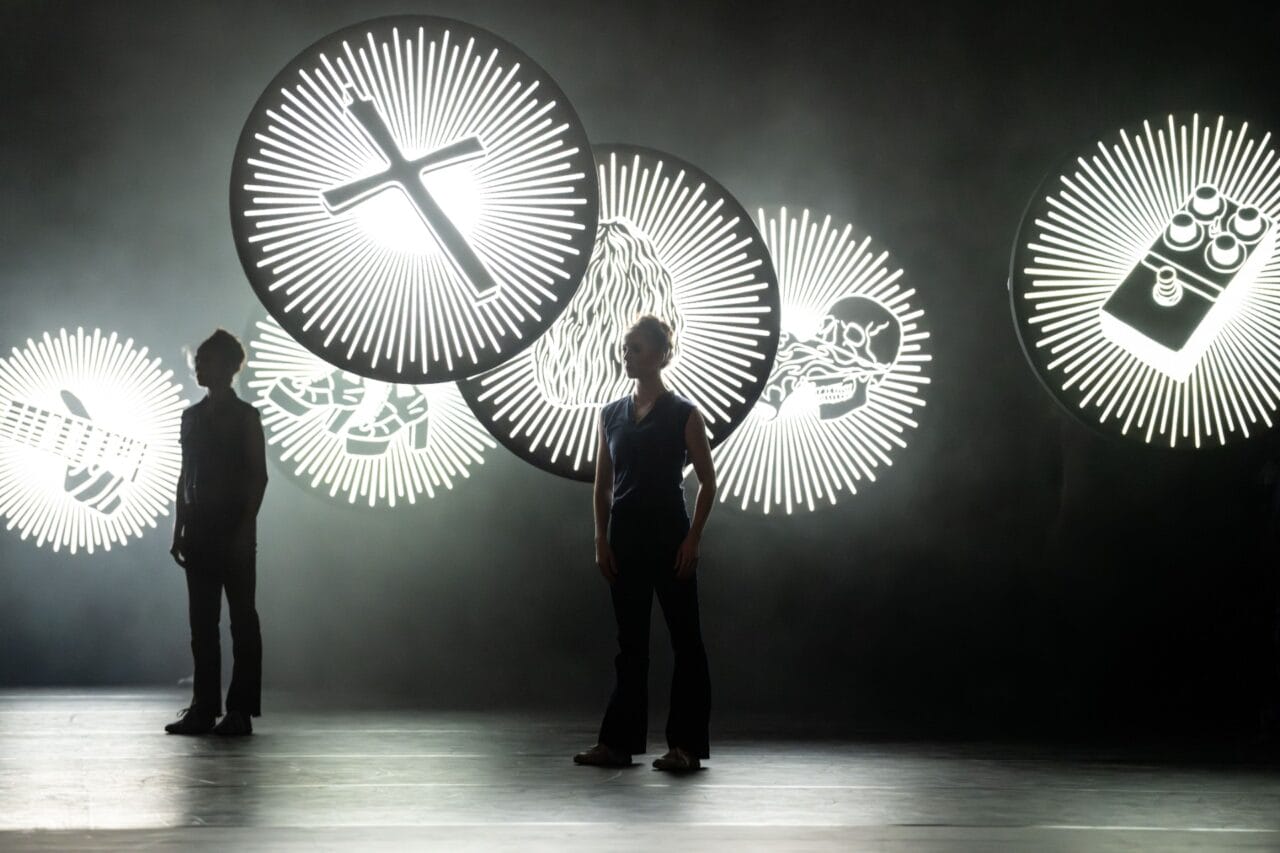 Two people stand onstage in front of large circular illuminated signs featuring symbols including a cross, a beard, a snake, and a LEGO brick, with a dark and smoky background.