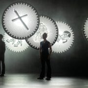 Two people stand onstage in front of large circular illuminated signs featuring symbols including a cross, a beard, a snake, and a LEGO brick, with a dark and smoky background.