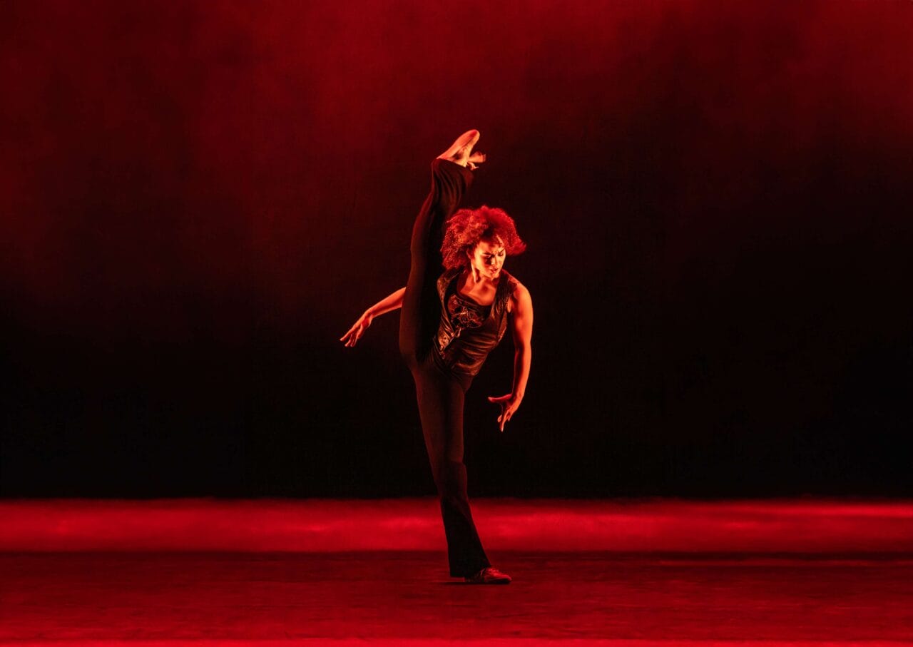 A dancer in black clothing performs a high leg kick on stage, with dramatic red lighting and a dark background.