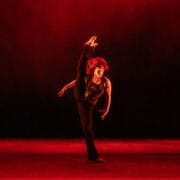 A dancer in black clothing performs a high leg kick on stage, with dramatic red lighting and a dark background.