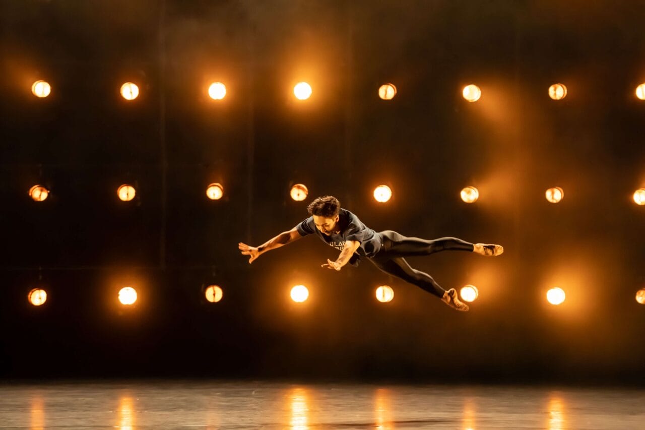 A dancer is captured mid-air in a leap on stage, with a grid of bright lights illuminating the dark background.