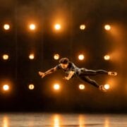 A dancer is captured mid-air in a leap on stage, with a grid of bright lights illuminating the dark background.