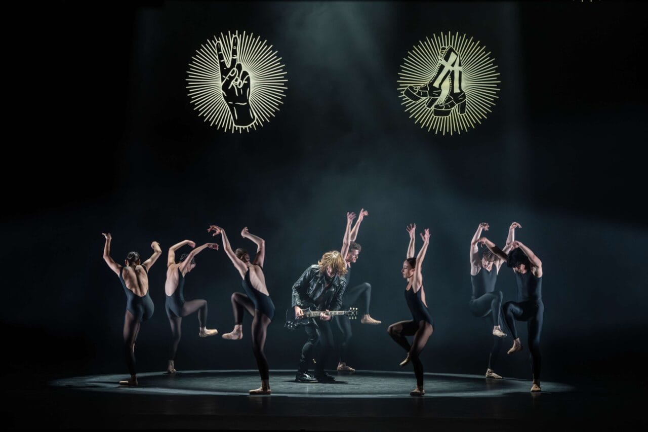 A group of ballet dancers performs on stage around a guitarist in a leather jacket, with symbolic illuminated graphics overhead.