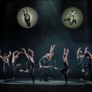 A group of ballet dancers performs on stage around a guitarist in a leather jacket, with symbolic illuminated graphics overhead.