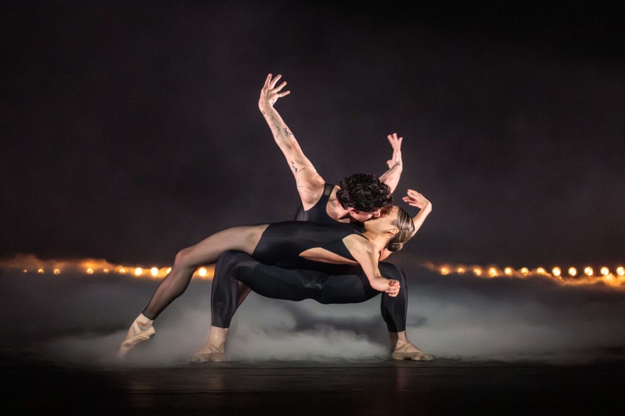 Two ballet dancers in black costumes perform an expressive, dramatic pose on a misty stage with a row of lights in the background.