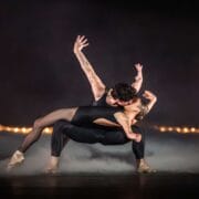 Two ballet dancers in black costumes perform an expressive, dramatic pose on a misty stage with a row of lights in the background.