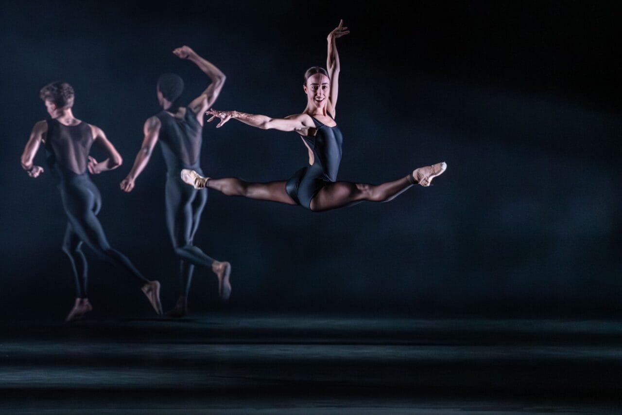 A ballet dancer performs a mid-air split leap in front of two other dancers on a dimly lit stage, all wearing dark leotards.