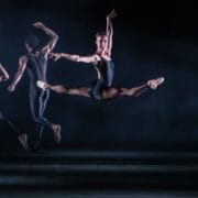 A ballet dancer performs a mid-air split leap in front of two other dancers on a dimly lit stage, all wearing dark leotards.