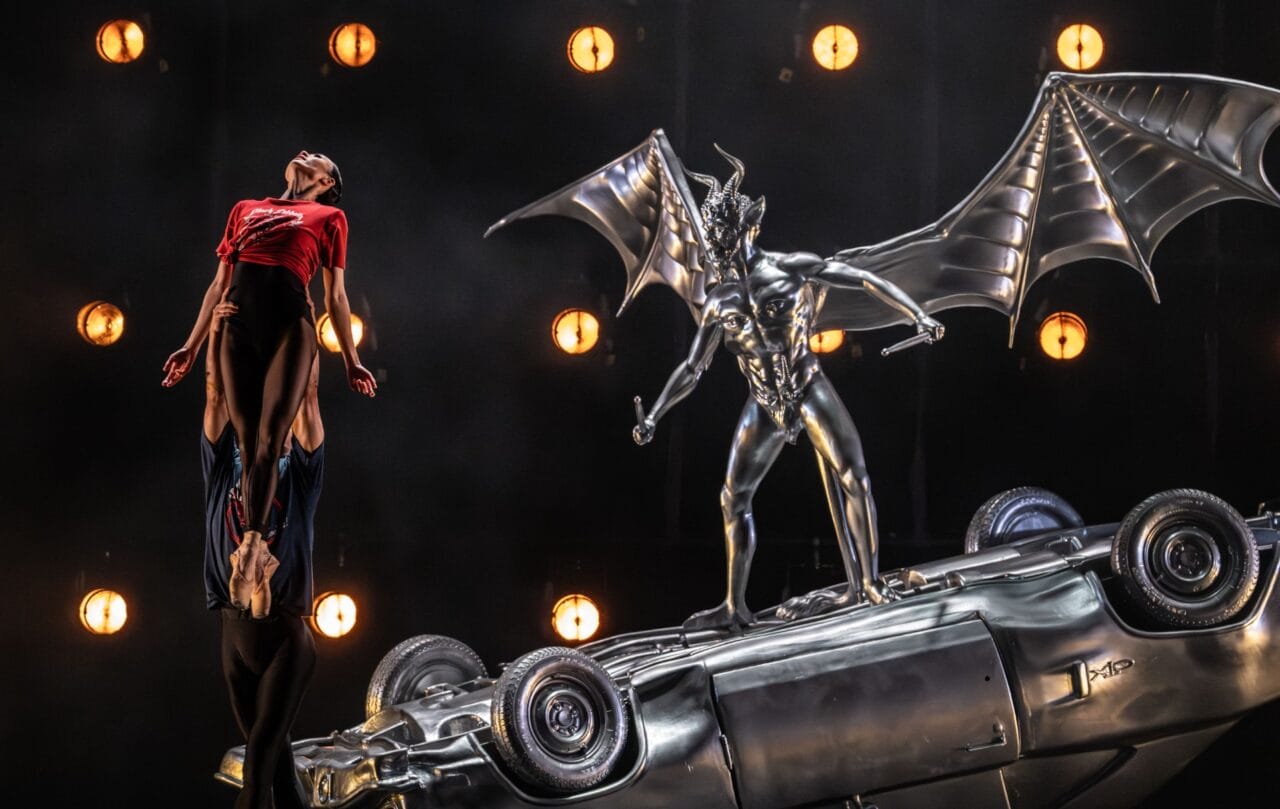 A performer is lifted into the air beside a metallic winged demon statue standing on a flipped car, with stage lights in the background.