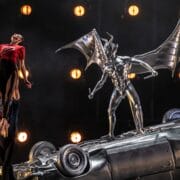 A performer is lifted into the air beside a metallic winged demon statue standing on a flipped car, with stage lights in the background.