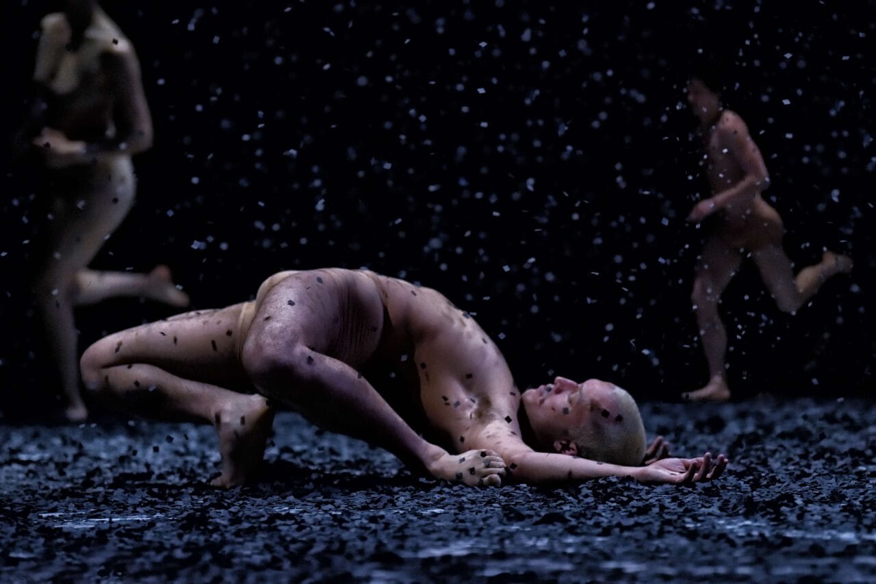 A person lies curled on the ground while others move in the background amid falling debris in a dark setting.