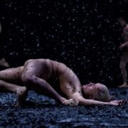 A person lies curled on the ground while others move in the background amid falling debris in a dark setting.