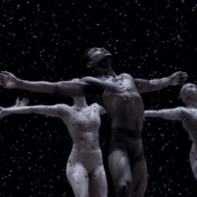 Four dancers with arms outstretched perform under artificial snow on a dark stage, with one dancer in front wearing dark attire and the others in lighter costumes.