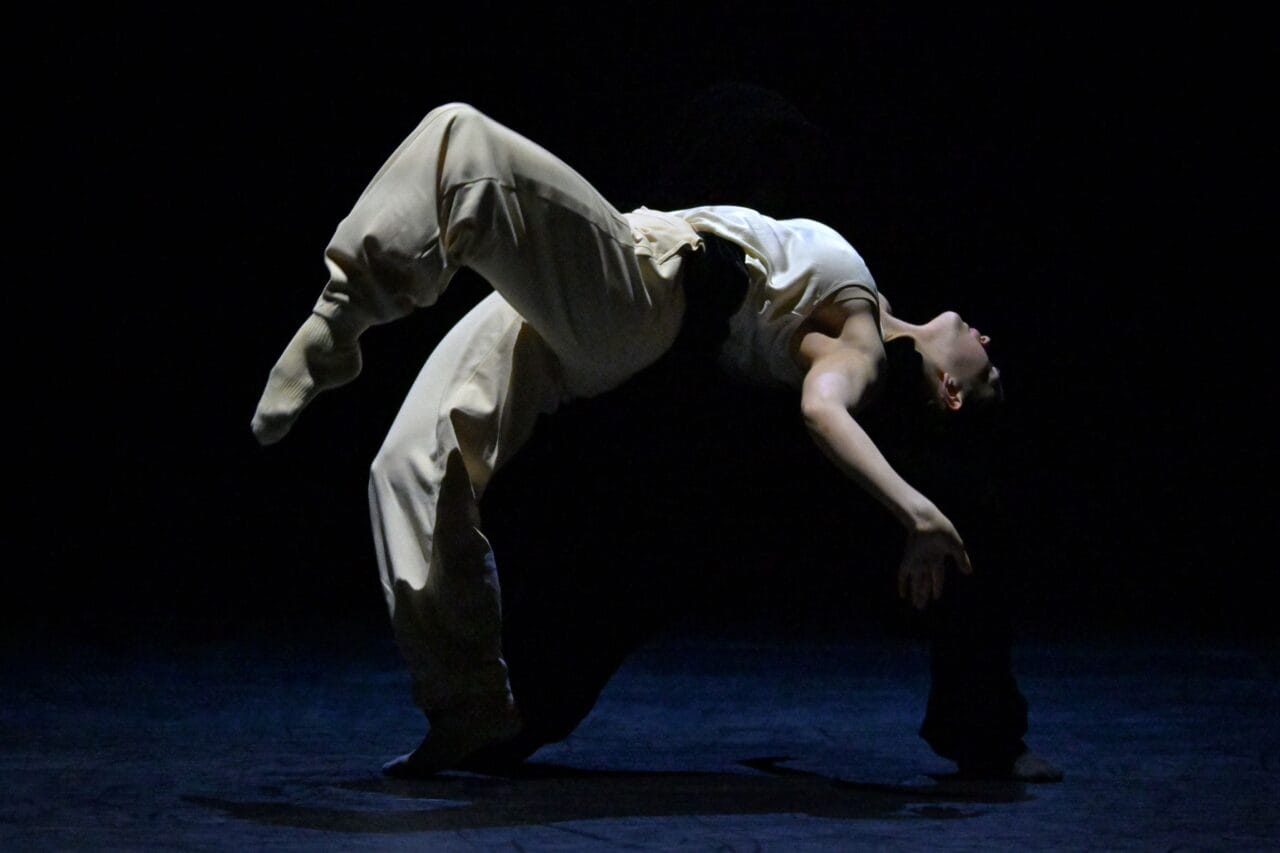 A person in loose pants and a tank top performs an arched backbend dance move on a dark stage, with dramatic lighting highlighting their form.