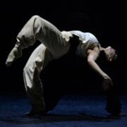 A person in loose pants and a tank top performs an arched backbend dance move on a dark stage, with dramatic lighting highlighting their form.