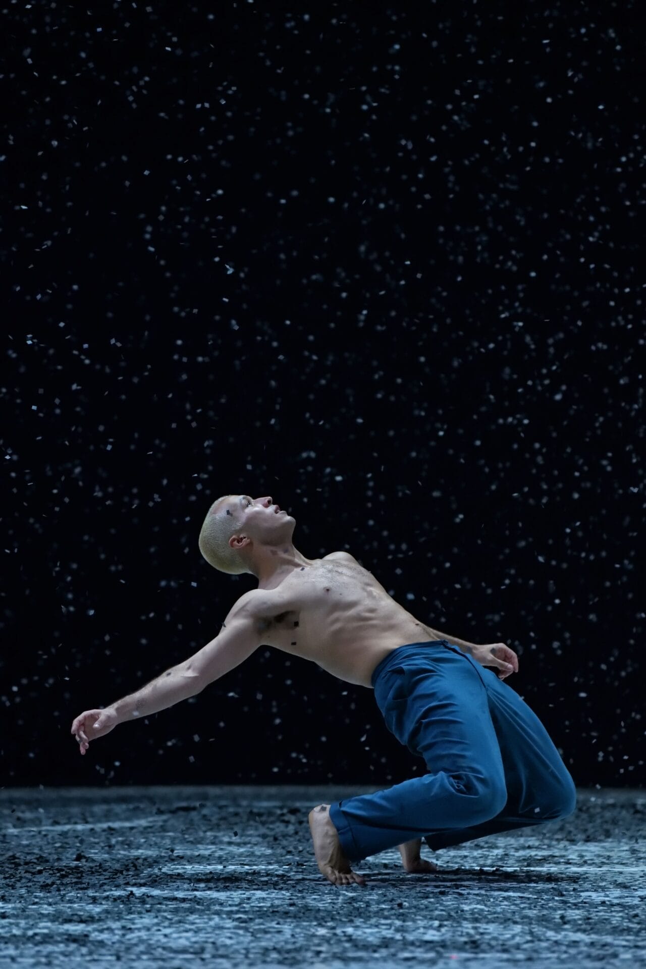 A shirtless person with short blond hair, wearing blue pants, leans back dramatically on a dark stage with falling water or snow.