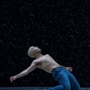 A shirtless person with short blond hair, wearing blue pants, leans back dramatically on a dark stage with falling water or snow.
