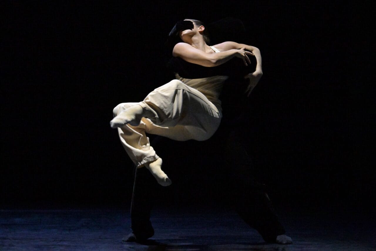 Two dancers perform on a dark stage; one dancer lifts and embraces the other, who wears light-colored clothing, creating a dramatic contrast.