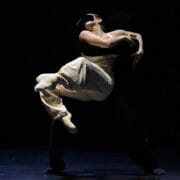 Two dancers perform on a dark stage; one dancer lifts and embraces the other, who wears light-colored clothing, creating a dramatic contrast.