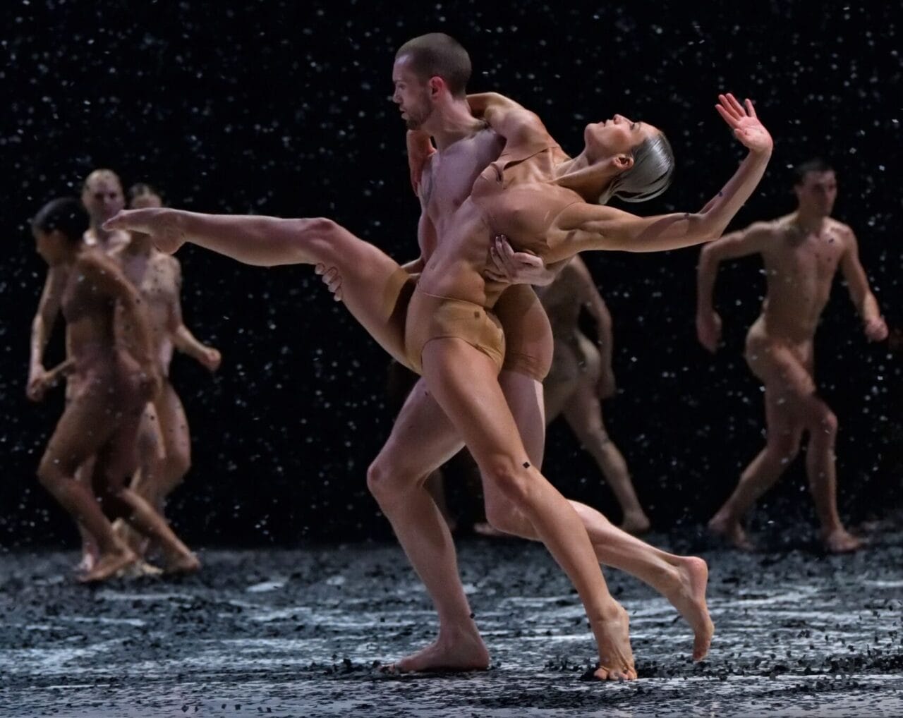 A group of dancers perform on stage under falling artificial snow, with two central figures in a dramatic dance pose.