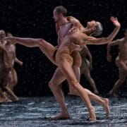 A group of dancers perform on stage under falling artificial snow, with two central figures in a dramatic dance pose.