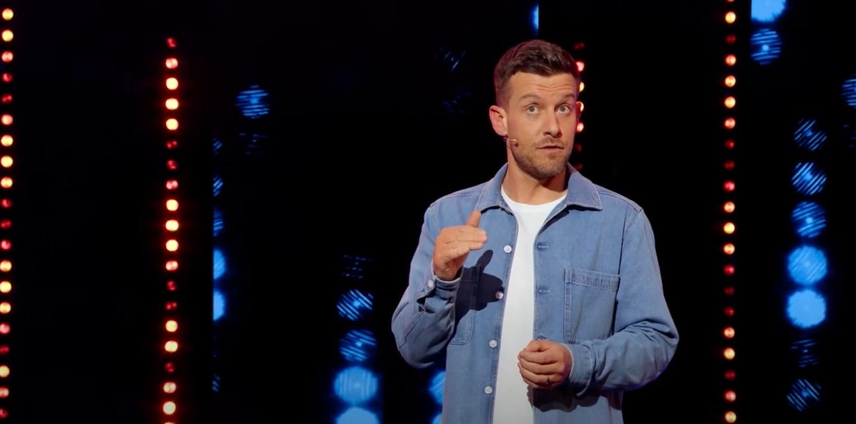 A man in a denim jacket stands on stage speaking, with vertical rows of lights in the background.