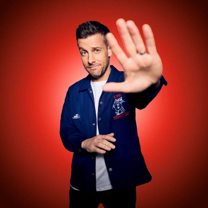 A man in a blue jacket stands against a red background with his right hand raised toward the camera, as if gesturing to stop.