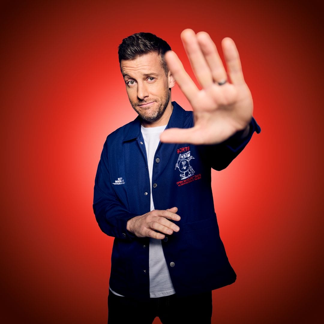 A man in a blue jacket stands against a red background with his right hand raised toward the camera, as if gesturing to stop.