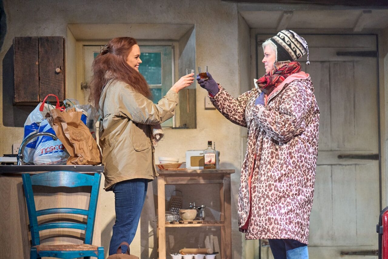 Two women in winter clothing clink glasses in a rustic kitchen; shopping bags and a bottle of whiskey are on the table beside them.