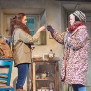 Two women in winter clothing clink glasses in a rustic kitchen; shopping bags and a bottle of whiskey are on the table beside them.