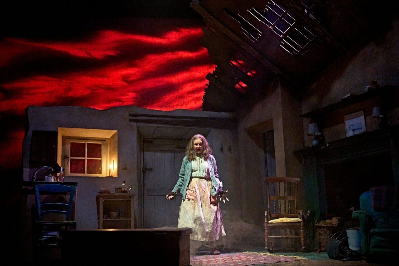 A woman stands center stage in a dimly lit, damaged room with red clouds projected behind her, wearing a cardigan and long skirt, her hands held in front of her.