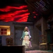 A woman stands center stage in a dimly lit, damaged room with red clouds projected behind her, wearing a cardigan and long skirt, her hands held in front of her.