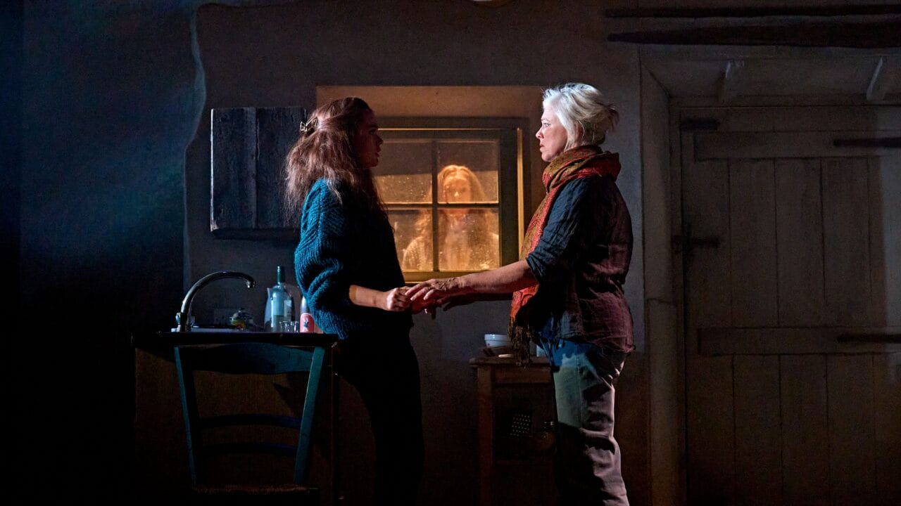 Two women stand holding hands in a dimly lit room, while another person looks in through the window from outside.