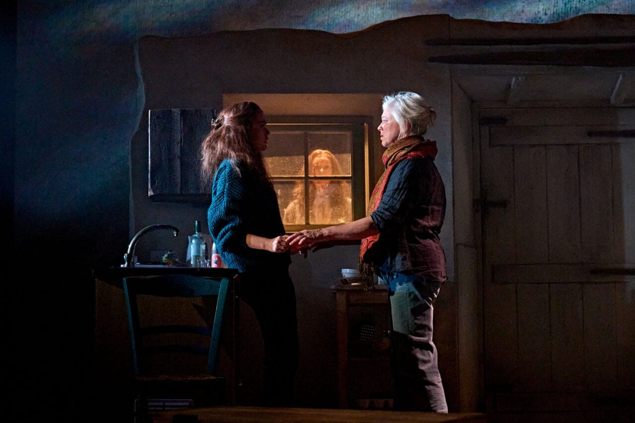 Two women stand holding hands in a dimly lit room, while another person looks in through the window from outside.