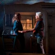 Two women stand holding hands in a dimly lit room, while another person looks in through the window from outside.