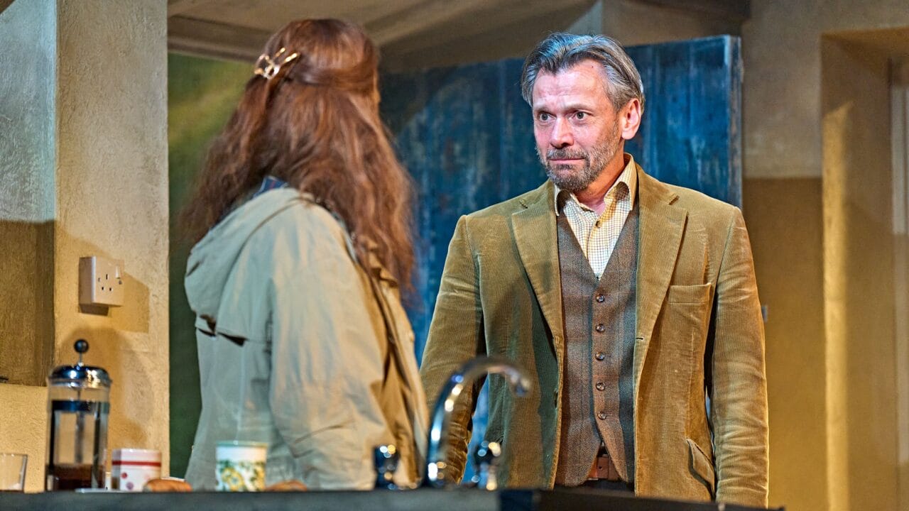 A man in a brown suit stands facing a woman with long hair in a kitchen, both appearing engaged in a serious conversation.