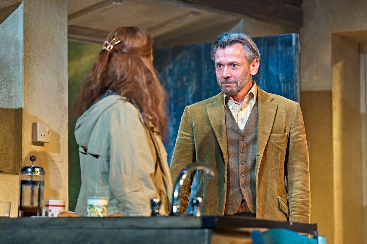 A man in a brown suit stands facing a woman with long hair in a kitchen, both appearing engaged in a serious conversation.