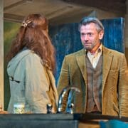 A man in a brown suit stands facing a woman with long hair in a kitchen, both appearing engaged in a serious conversation.