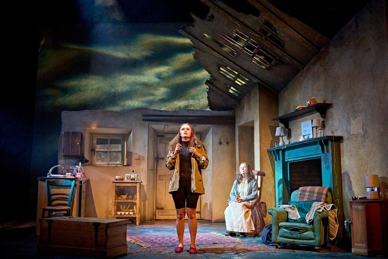 A woman stands speaking at the front of a rundown room while another woman sits in an armchair; the set shows a dilapidated ceiling and worn furnishings under stage lighting.