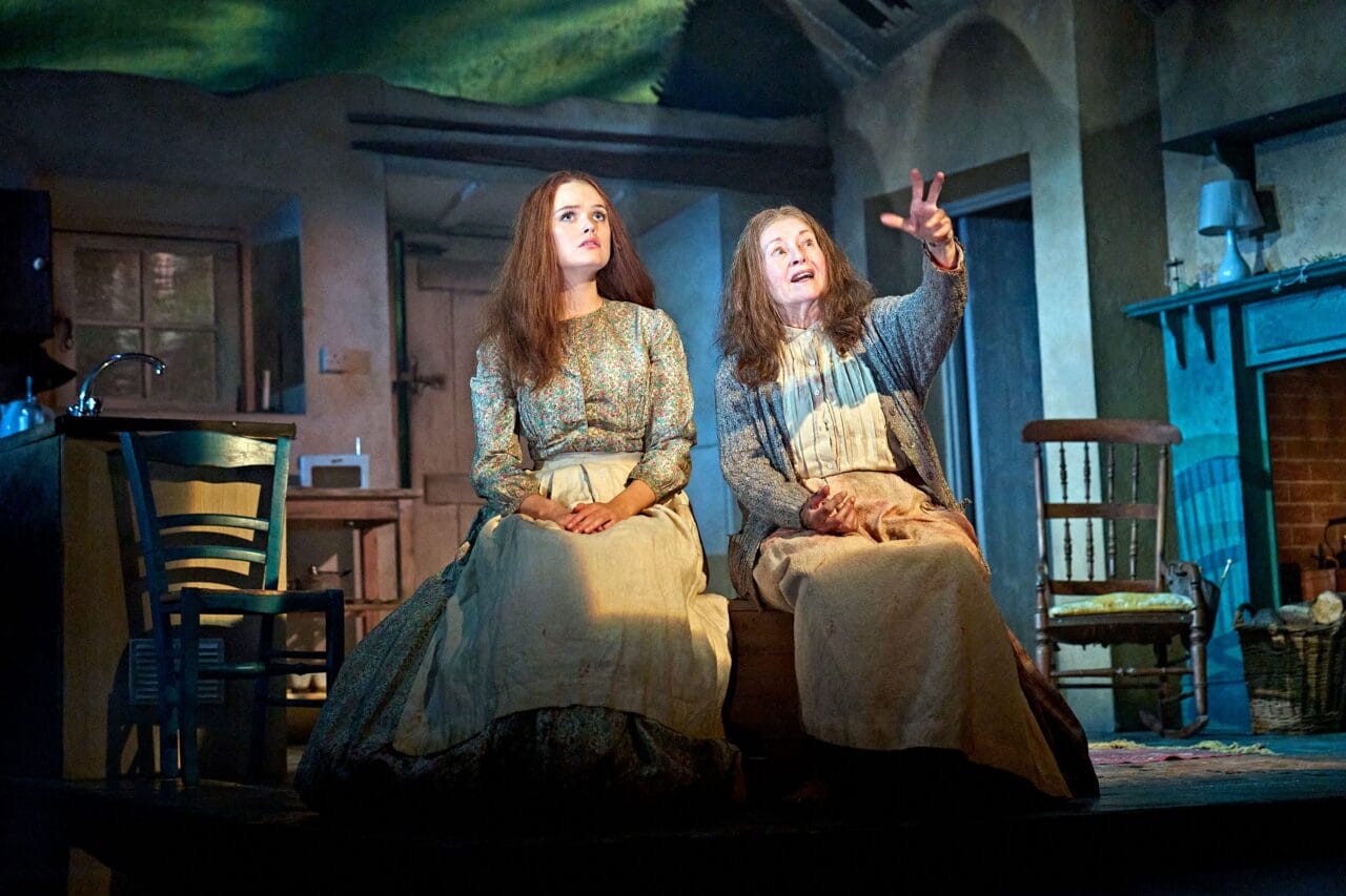 Two women in period costumes sit on a wooden box in a rustic, dimly lit room; one gestures animatedly while the other looks ahead.