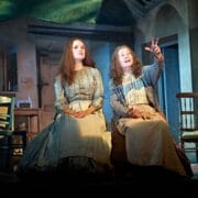 Two women in period costumes sit on a wooden box in a rustic, dimly lit room; one gestures animatedly while the other looks ahead.