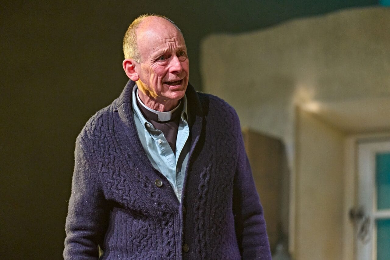 An older man wearing a clerical collar and knitted sweater stands indoors, appearing emotional with a distressed facial expression.