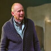 An older man wearing a clerical collar and knitted sweater stands indoors, appearing emotional with a distressed facial expression.