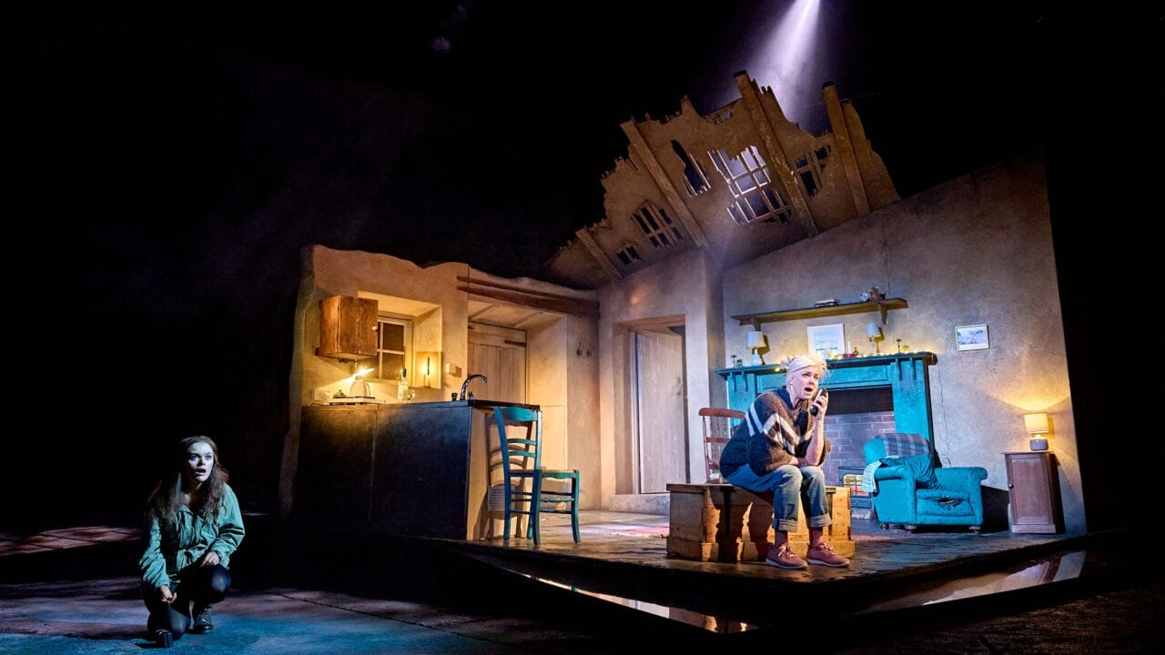 A stage scene shows a woman crouching in darkness at left and an older man sitting under a spotlight in a dilapidated, partially destroyed room set with visible kitchen and living area.