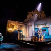 A stage scene shows a woman crouching in darkness at left and an older man sitting under a spotlight in a dilapidated, partially destroyed room set with visible kitchen and living area.
