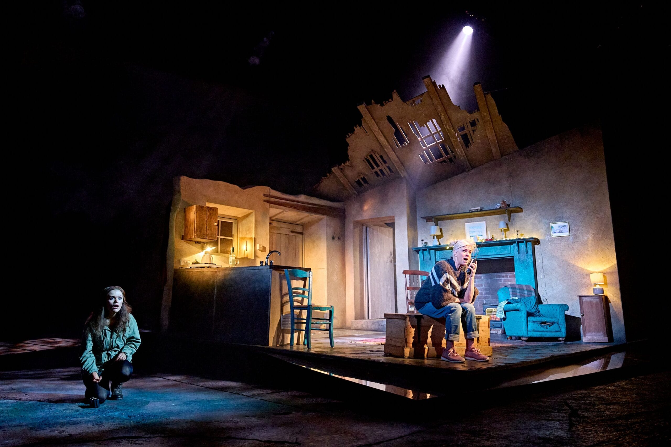 A stage scene shows a woman crouching in darkness at left and an older man sitting under a spotlight in a dilapidated, partially destroyed room set with visible kitchen and living area.
