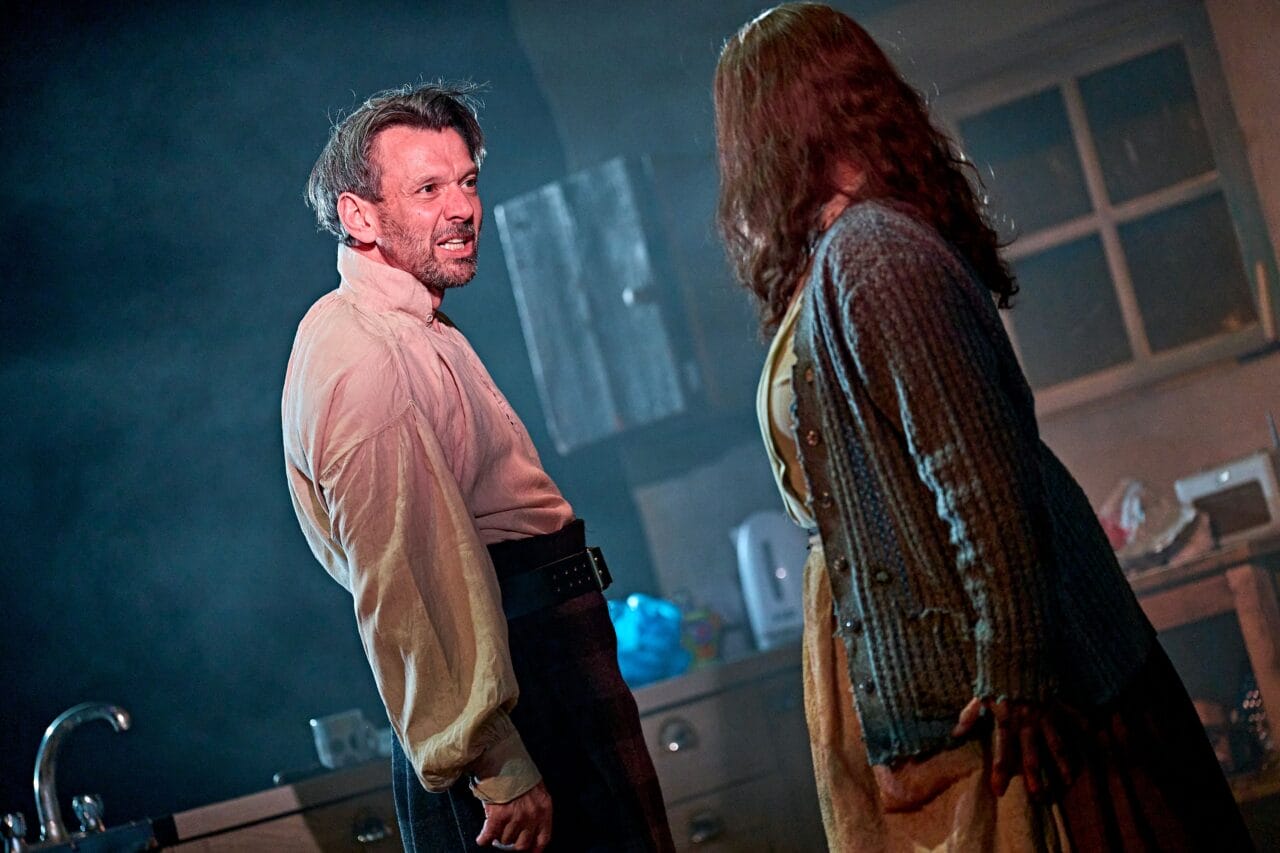 A man and woman stand facing each other in a dimly lit kitchen, with the man looking tense and the woman turned slightly away.